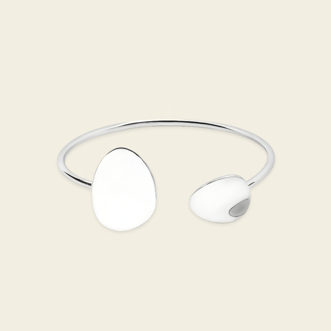 A minimalist silver-toned open cuff bangle featuring two smooth, organic-shaped disc elements, displayed on a solid light beige background.