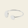 A minimalist silver open cuff bracelet with two flat, oval disc charms, displayed on a light cream background.
