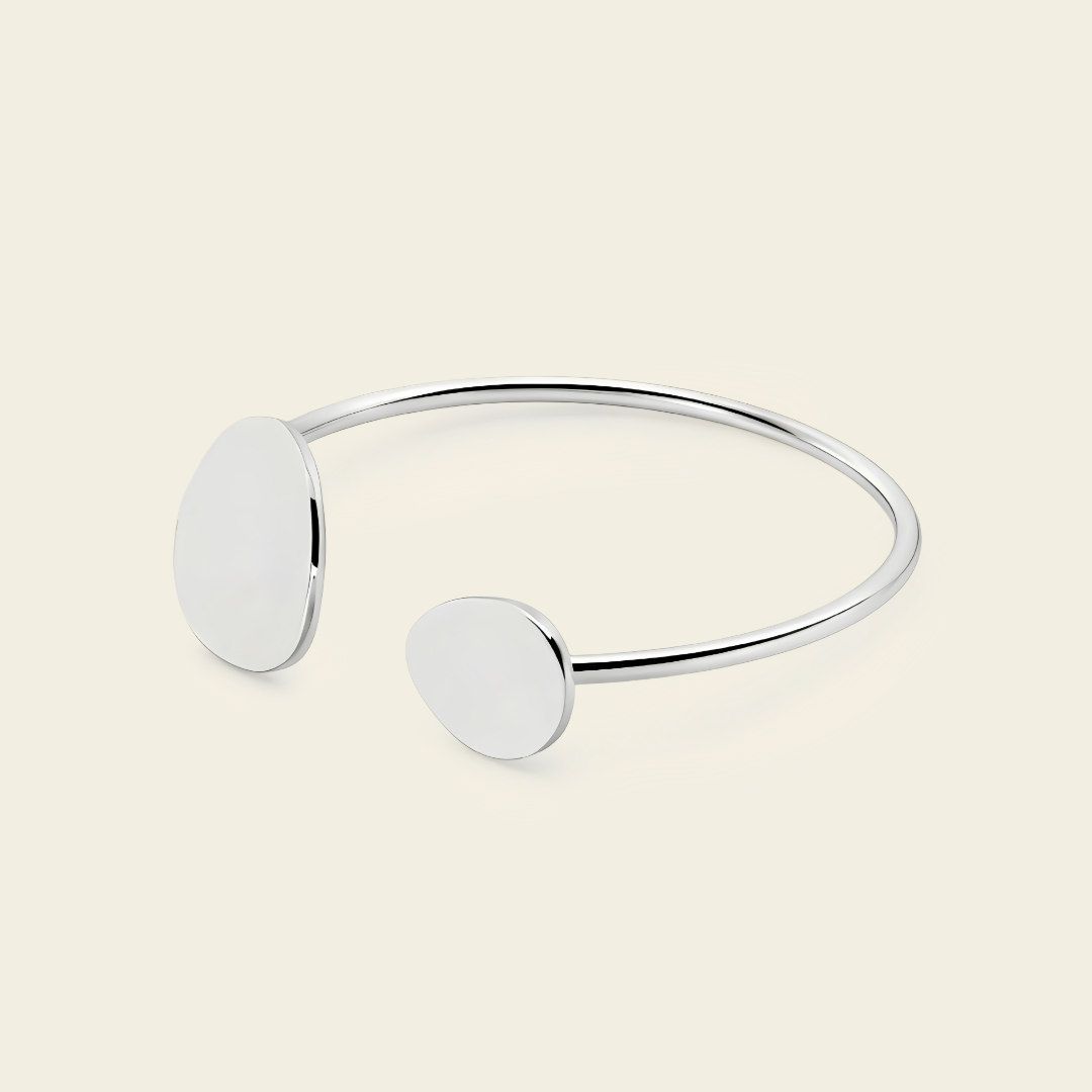 A minimalist silver open cuff bracelet with two flat, oval disc charms, displayed on a light cream background.