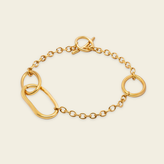 A gold chain bracelet featuring a large flat oval link, two smaller circular links, and a toggle clasp, displayed on a light cream background.