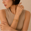 A close-up of a woman wearing a chunky gold chain-link bracelet with interlocking oval links, styled with a beige ribbed sleeveless top against a warm beige background.