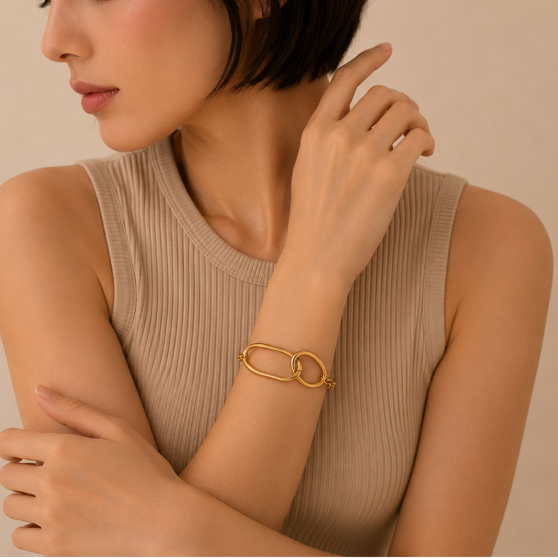 A close-up of a woman wearing a chunky gold chain-link bracelet with interlocking oval links, styled with a beige ribbed sleeveless top against a warm beige background.