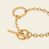 Gold-tone toggle ring bracelet with a short chain extension and a flat toggle bar, shown on a light cream background.