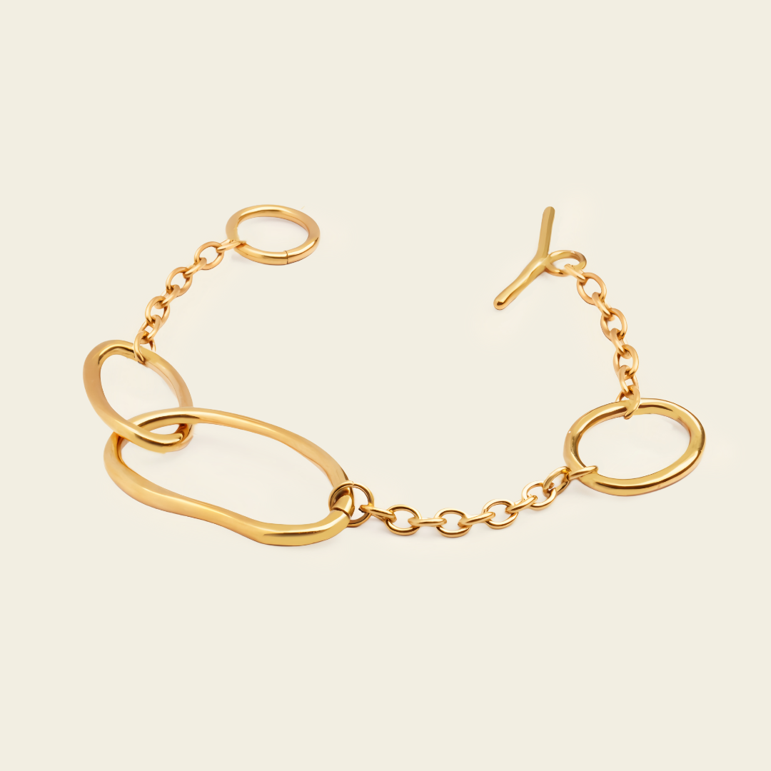 A gold-toned chain bracelet featuring large, organic wavy oval links, a smaller circular link, and a toggle clasp, displayed on a light cream background.