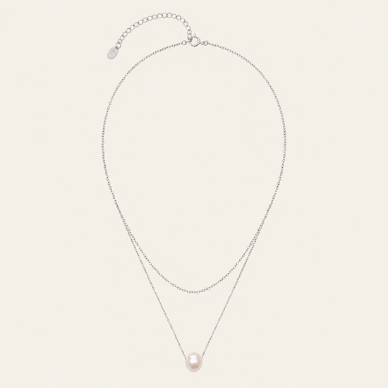 A delicate double-layered necklace featuring a silver-tone chain with an extender clasp and a single round, pale pink pearl pendant, displayed on a light cream background.