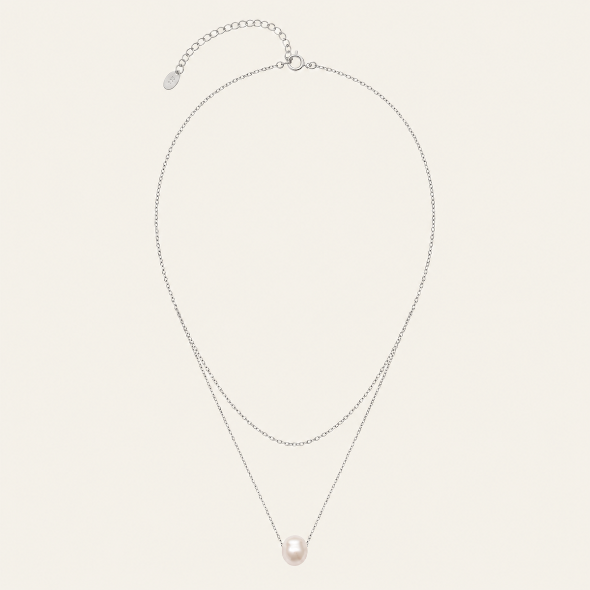 A delicate double-layered necklace featuring a silver-tone chain with an extender clasp and a single round, pale pink pearl pendant, displayed on a light cream background.