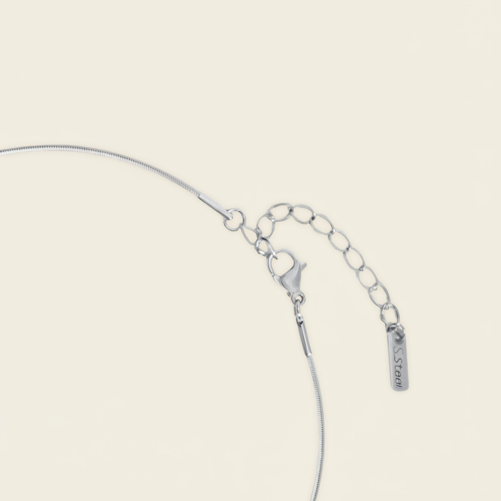 Close-up of a silver chain necklace with a lobster clasp, extension chain, and a small rectangular tag on a light cream background.
