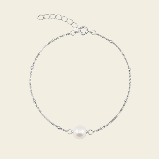 A delicate silver chain bracelet featuring a single white freshwater pearl at the center, displayed on a light beige background.