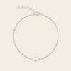 A delicate silver chain bracelet featuring a single white freshwater pearl at the center, displayed on a light beige background.