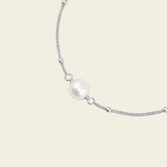 A delicate silver chain bracelet featuring a single lustrous white pearl bead at its center, displayed on a solid light beige background.
