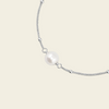 A delicate silver chain bracelet featuring a single lustrous white pearl bead at its center, displayed on a solid light beige background.