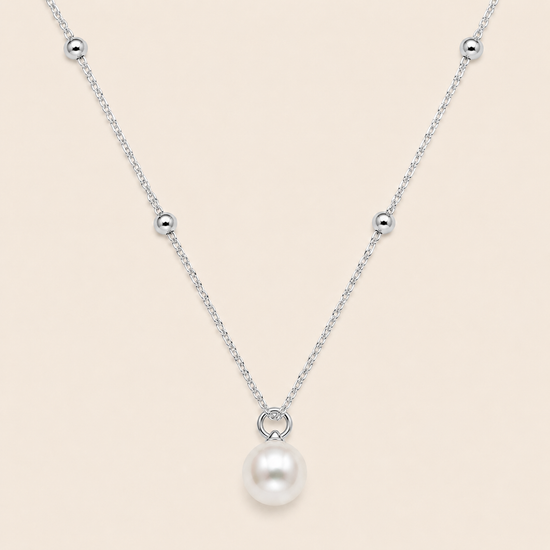 A delicate silver chain necklace featuring a single white pearl pendant suspended from a small silver ring, accented with small silver beads along the chain, set against a solid pale beige background.