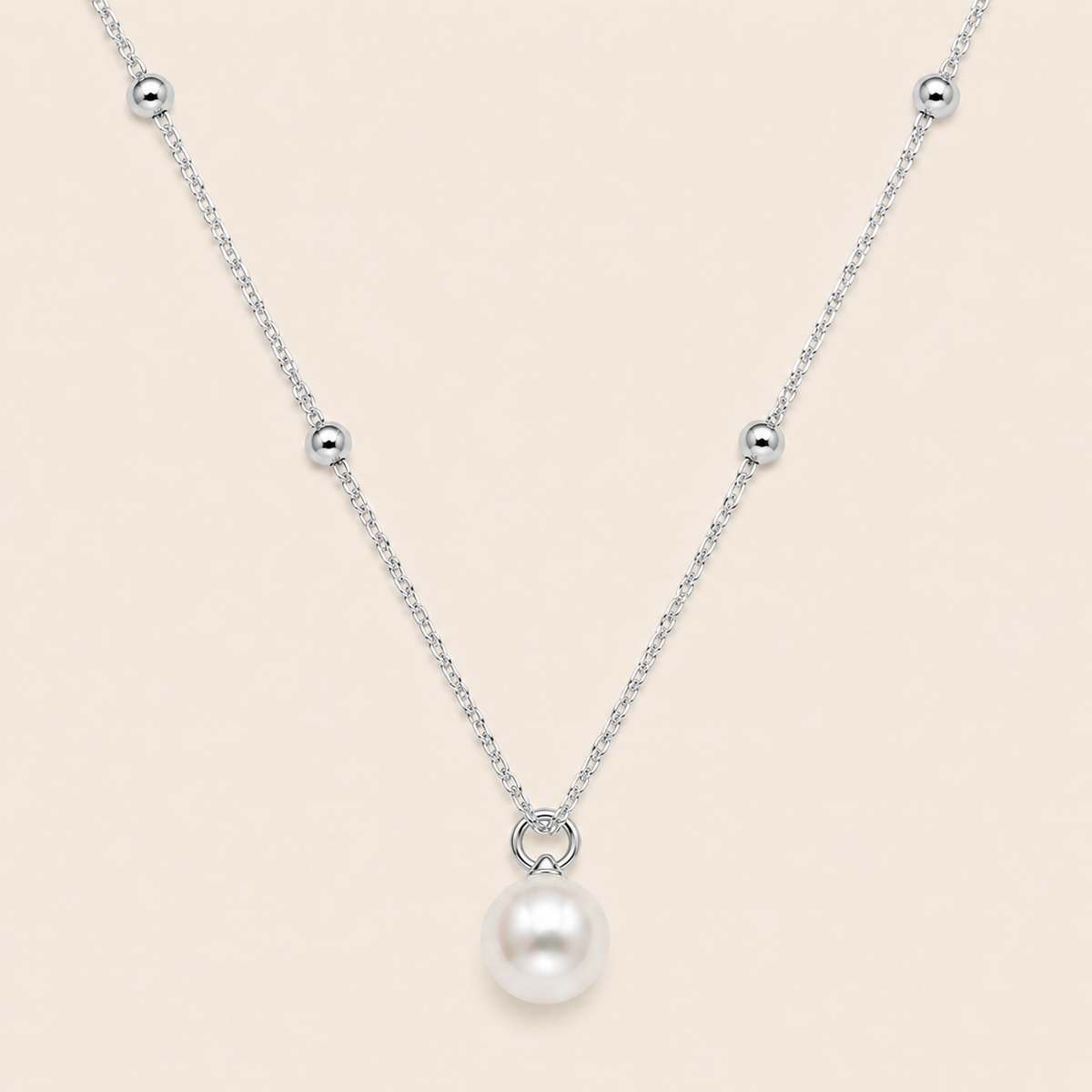 A delicate silver chain necklace featuring a single white pearl pendant suspended from a small silver ring, accented with small silver beads along the chain, set against a solid pale beige background.