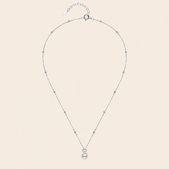 A delicate silver chain necklace featuring a single white pearl pendant, accented with small sparkling beads spaced along the chain, displayed against a solid pale beige background.