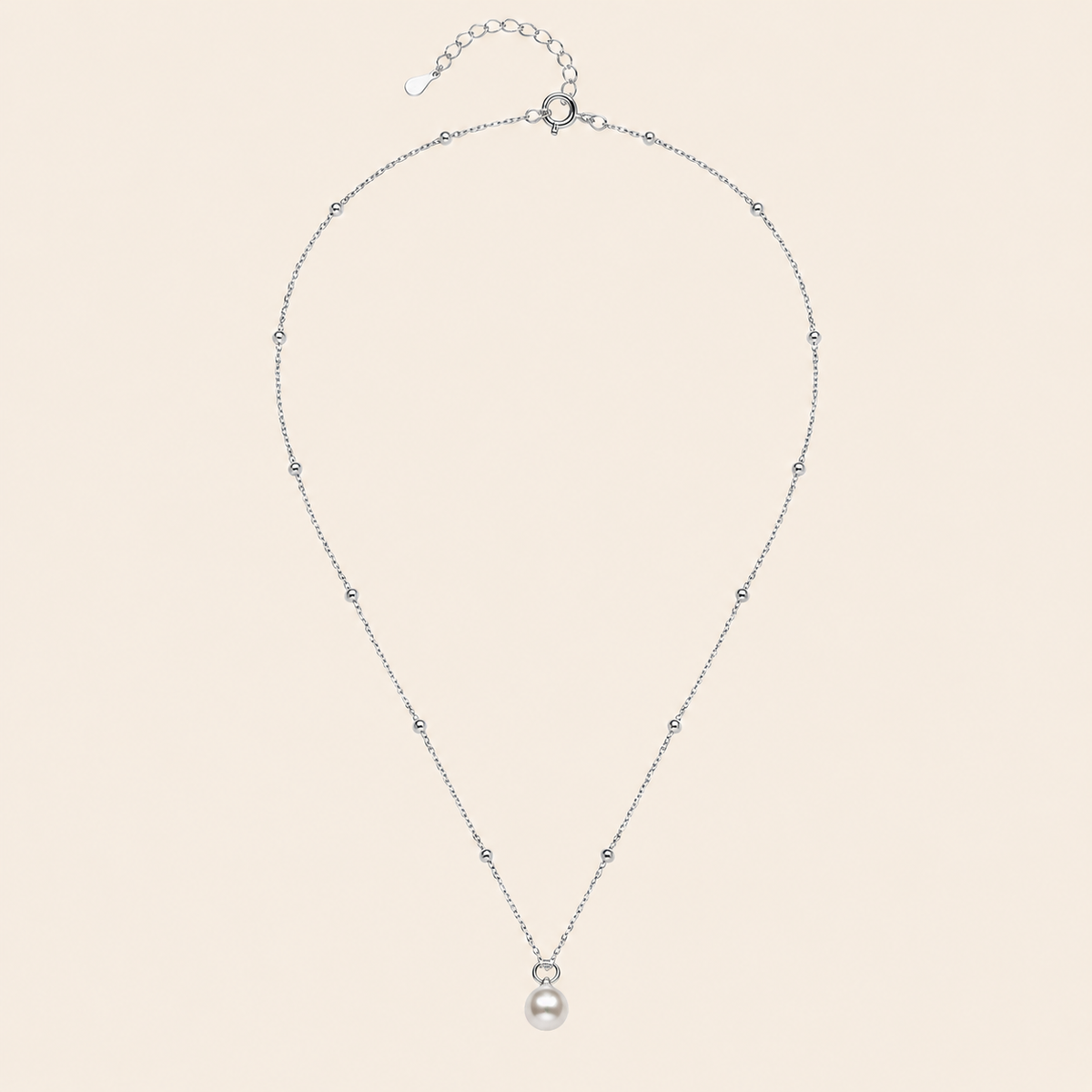 A delicate silver chain necklace featuring a single white pearl pendant, accented with small sparkling beads spaced along the chain, displayed against a solid pale beige background.