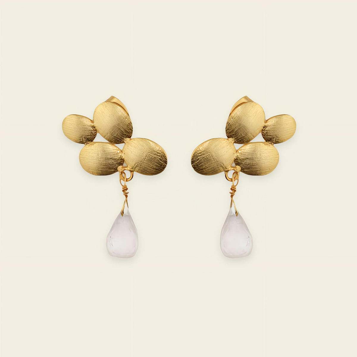 Gold earrings with leaf-like designs and white teardrop-shaped stones on a beige background