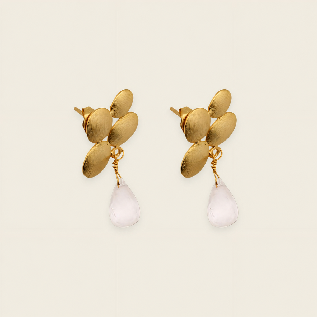 Gold earrings with leaf-like designs and white teardrop-shaped stones on a beige background