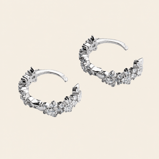 A pair of silver-toned crescent-shaped hoop earrings adorned with clear cubic zirconia stones, displayed on a solid light beige background.