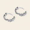 A pair of silver-toned crescent-shaped hoop earrings adorned with clear cubic zirconia stones, displayed on a solid light beige background.