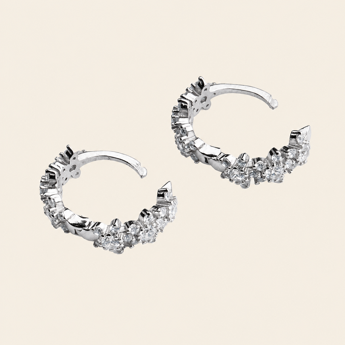 A pair of silver-toned crescent-shaped hoop earrings adorned with clear cubic zirconia stones, displayed on a solid light beige background.