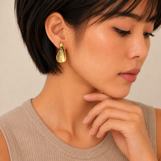 A close-up of a woman with short dark hair wearing a polished gold teardrop-shaped earring, paired with a beige ribbed tank top against a soft beige background.