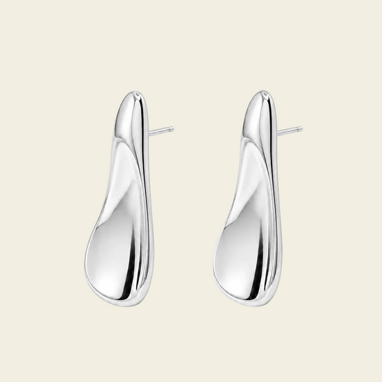 A pair of polished silver drop earrings with a smooth, organic teardrop shape and post backs, displayed on a solid light beige background.