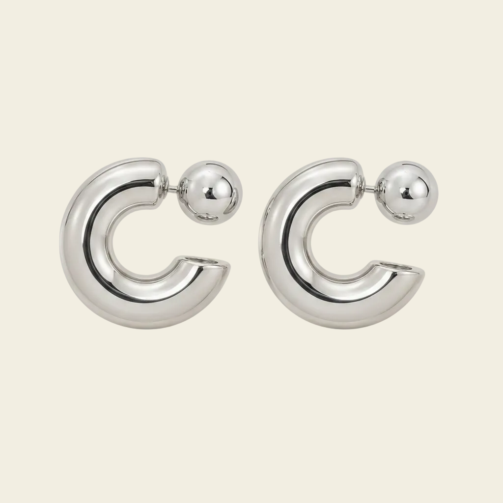 A pair of polished silver-toned hoop earrings with a small spherical stud attached at the front, shown on a plain warm off-white background.