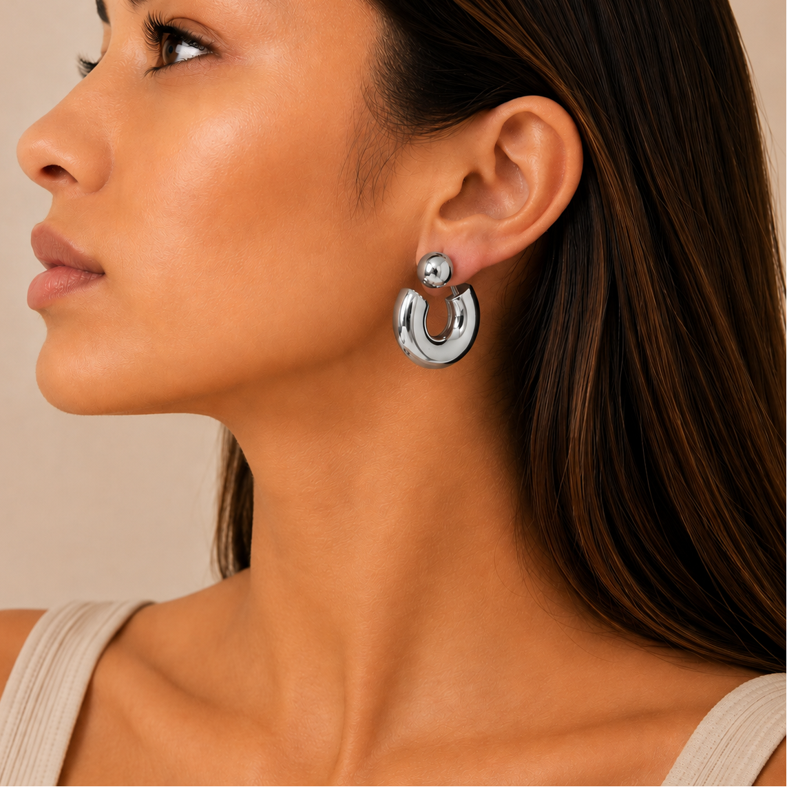 A close-up side profile of a woman wearing a chunky, polished silver-tone hoop earring with a small spherical stud above it.