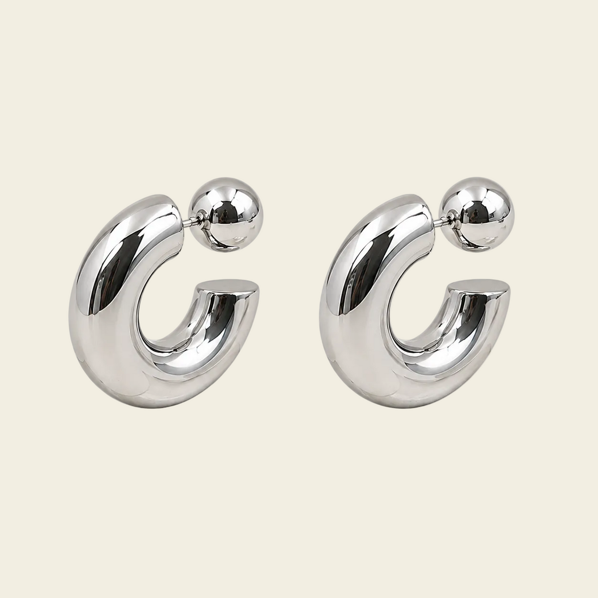 A pair of polished silver-toned hoop earrings with a small spherical bead at the top, displayed on a solid light beige background.