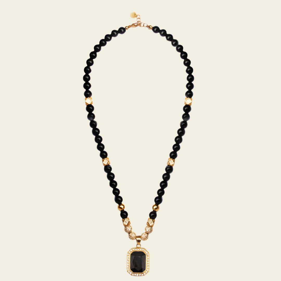 A beaded necklace featuring black round beads, small gold spacer beads, and a rectangular black pendant with a gold halo, displayed on a light beige background.