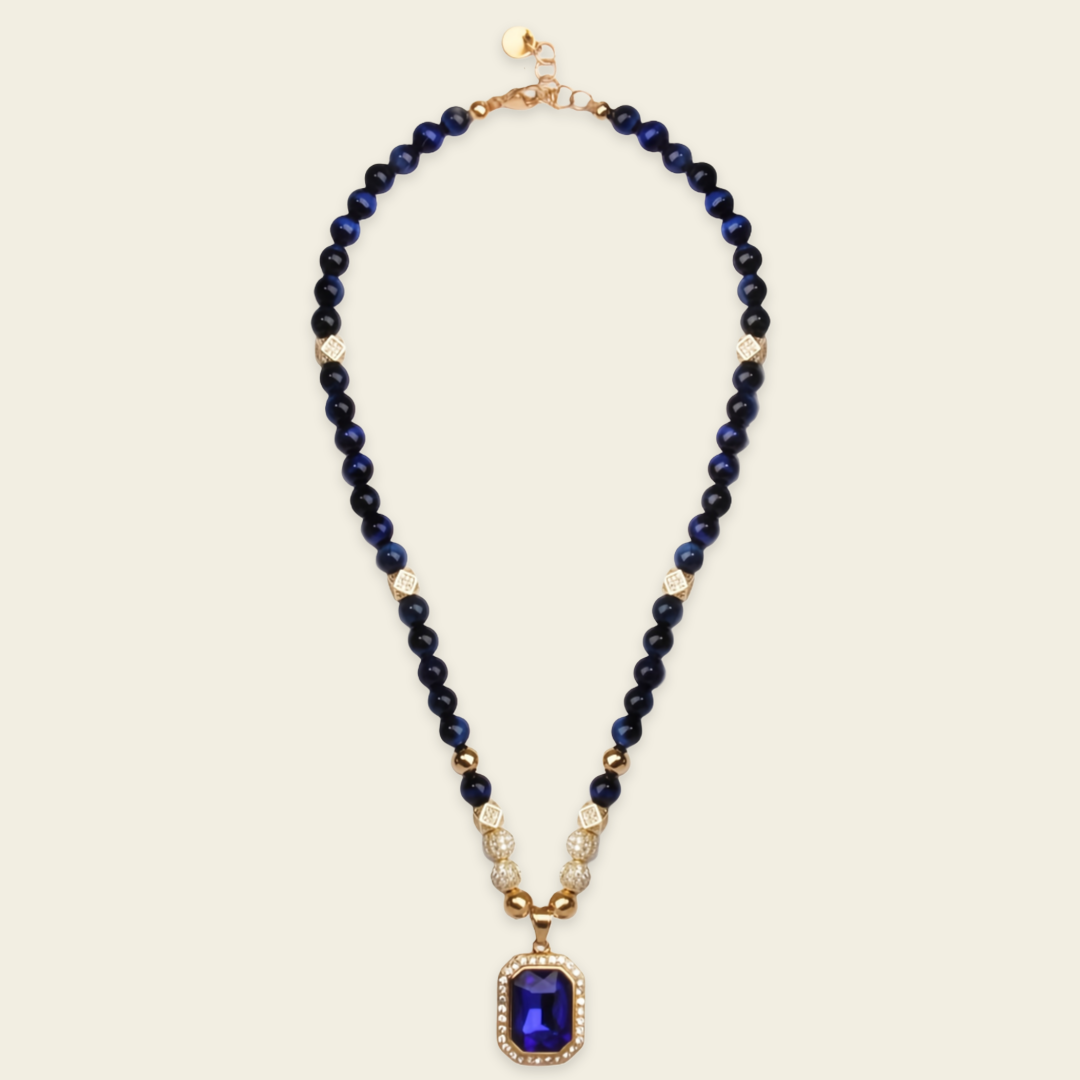 A beaded necklace featuring dark navy blue beads separated by small gold spacer beads, with a gold-toned rectangular pendant set with a deep blue faceted gemstone and tiny clear stones.
