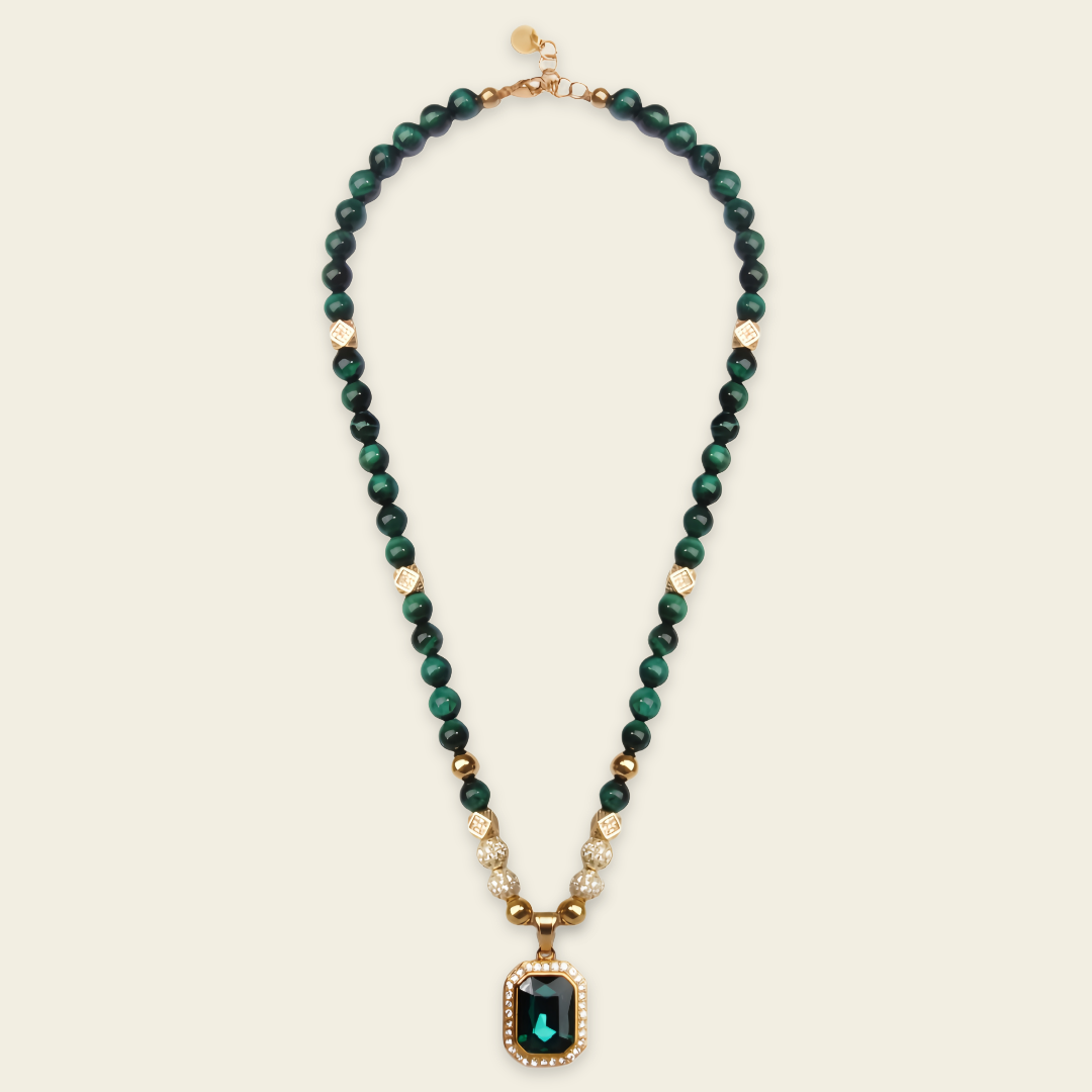 A beaded necklace featuring dark green beads, small gold spacer beads, and a central rectangular green gemstone pendant with a gold border, displayed on a light cream background.