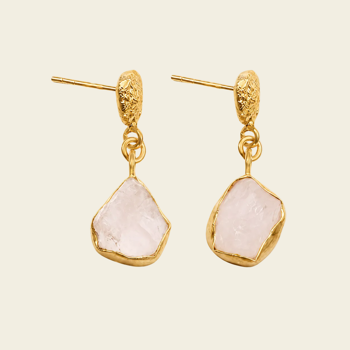 A pair of gold drop earrings featuring irregular, raw-cut pale pink stones, displayed against a solid light beige background.