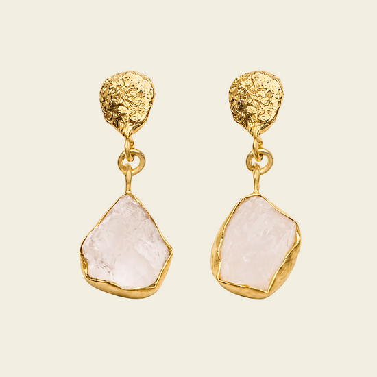 A pair of gold drop earrings featuring textured, organic-shaped gold tops and irregular pale pink stone pendants, displayed on a light cream background.