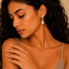 A close-up lifestyle shot of a woman with dark curly hair wearing a gold drop earring with a white teardrop-shaped stone, set against a warm beige background.