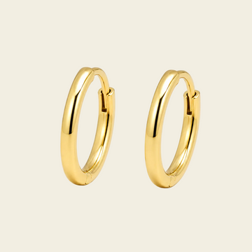 A pair of polished gold huggie hoop earrings on a warm cream background.