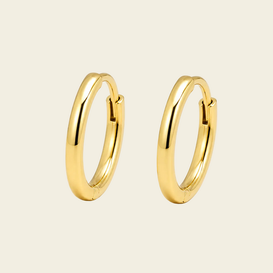 A pair of polished gold huggie hoop earrings on a warm cream background.
