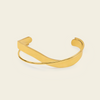 A polished gold-toned metal cuff bracelet with a smooth, curved design, displayed on a solid light beige background.