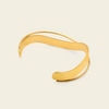 A polished gold-tone open cuff bracelet with a smooth, flowing wavy design, displayed on a light cream background.