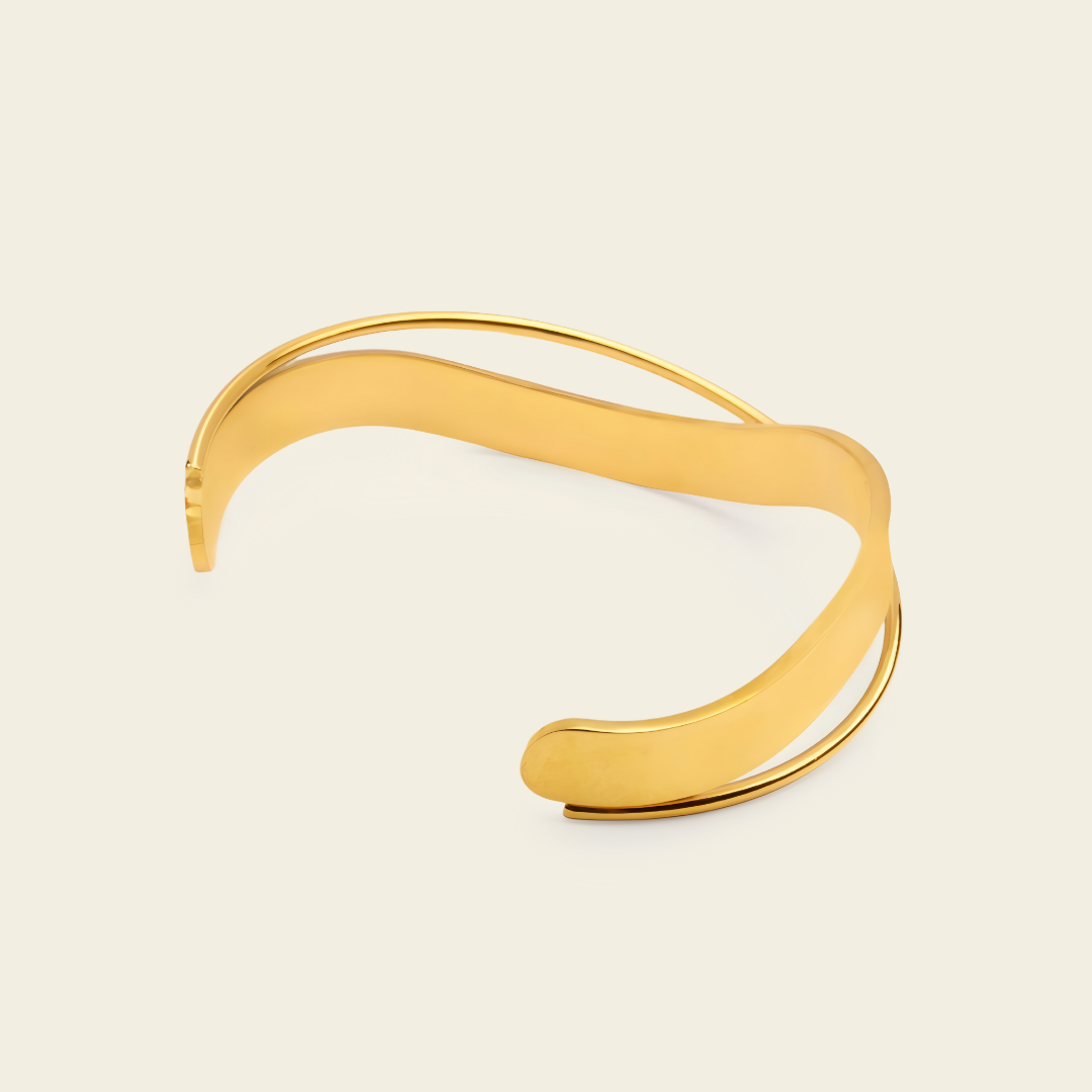 A polished gold-tone open cuff bracelet with a smooth, flowing wavy design, displayed on a light cream background.