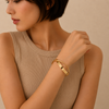 A woman wearing a slim gold bangle bracelet with a polished rectangular center detail, styled with a beige ribbed sleeveless top against a neutral beige background.