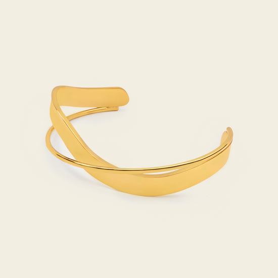 A polished gold-toned metal cuff bracelet with a smooth, curved open design, displayed on a solid light beige background.