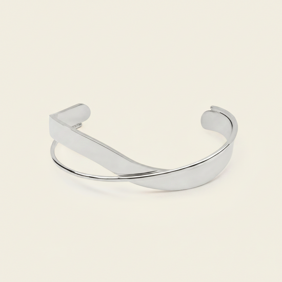 A polished silver-toned metal cuff bracelet with a smooth, curved design, displayed on a solid light beige background.