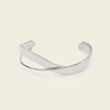 A polished silver-toned metal cuff bracelet with a smooth, curved design, displayed on a solid light beige background.