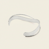 A polished silver-tone open cuff bracelet with a smooth, flowing wavy design, displayed on a light cream background.