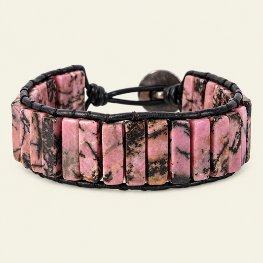 A handmade cuff bracelet made of rectangular pink rhodonite stones with black veining, wrapped in a black cord with a round metal toggle clasp, shown on a light cream background.