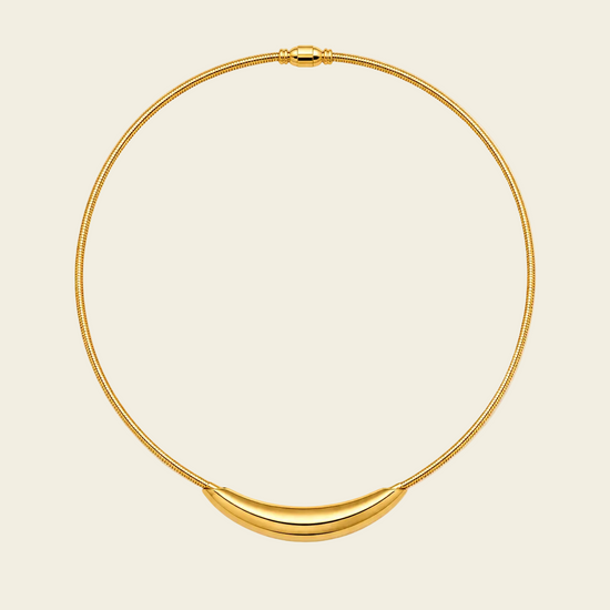 A gold snake-chain necklace with a polished crescent-shaped pendant, displayed on a light cream background.