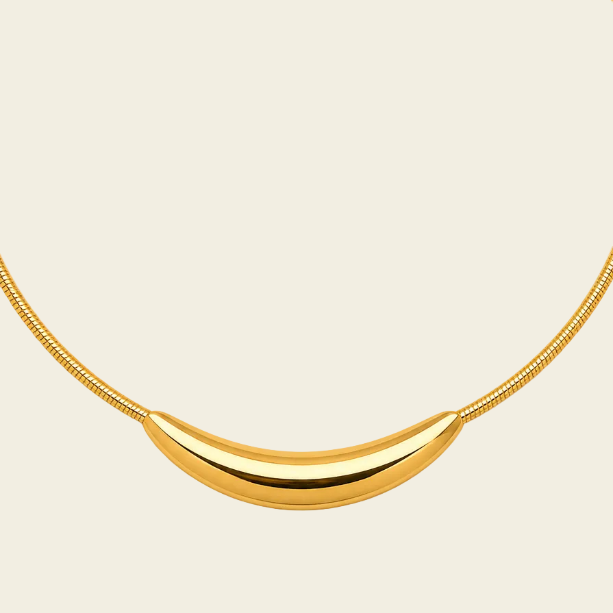 A gold necklace featuring a polished crescent-shaped pendant suspended from a textured snake chain, set against a solid cream background.
