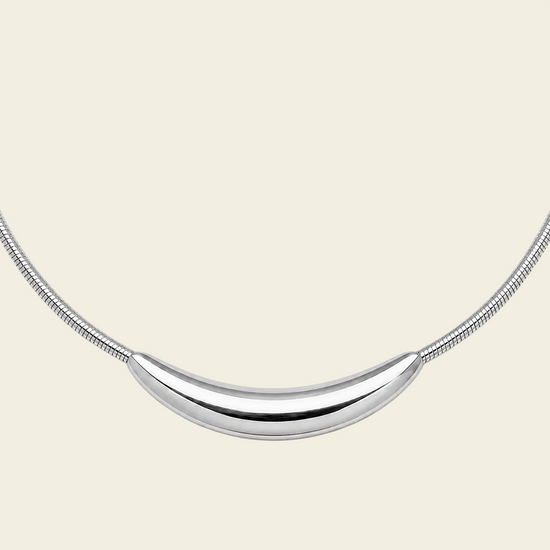 A silver crescent moon pendant necklace on a matching snake chain, displayed against a solid pale peach background.