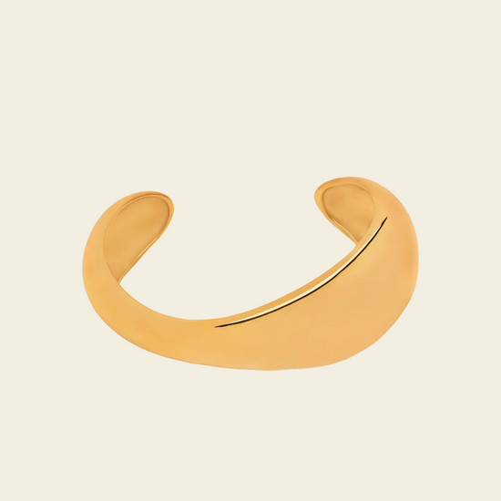 A minimalist, curved open cuff bracelet in a matte gold finish, displayed on a light cream background.